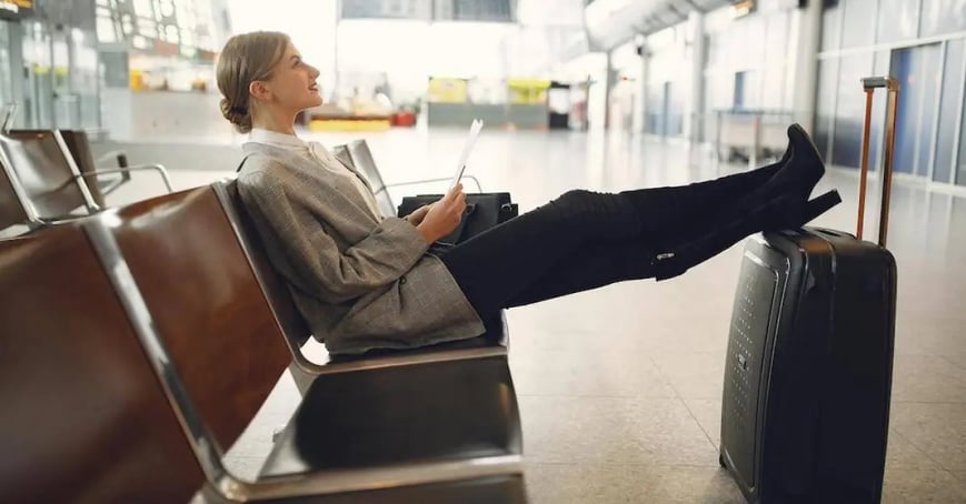 A woman sitting in an airport chair with her legs propped up on her suitcase