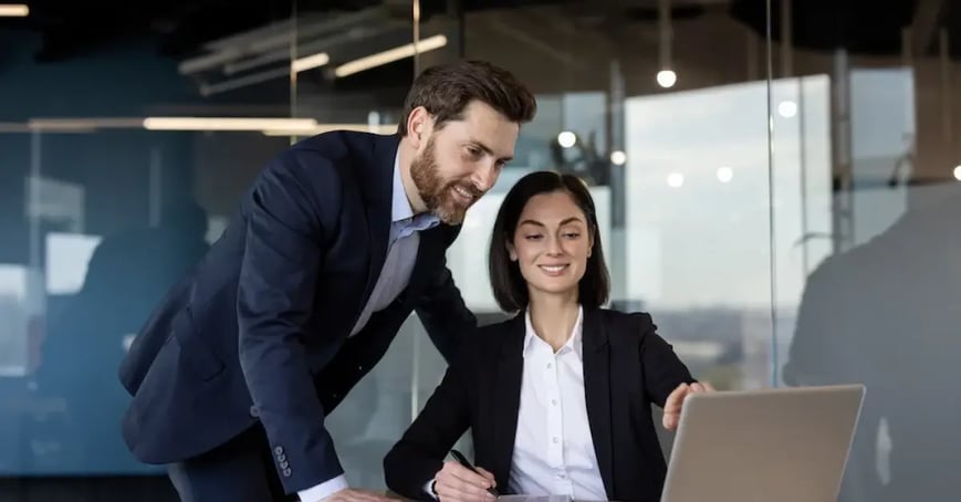 Two coworkers (one male, one female) wearing suits in an office building looking over the meeting objective on the laptop